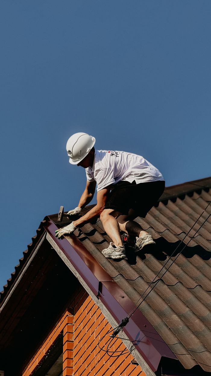 A roofer in a safety helmet renovating a brick house roof under clear blue skies.